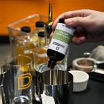 A bartender pours Durham Distillery’s cardamom bitters into a stainless steel cocktail shaker.